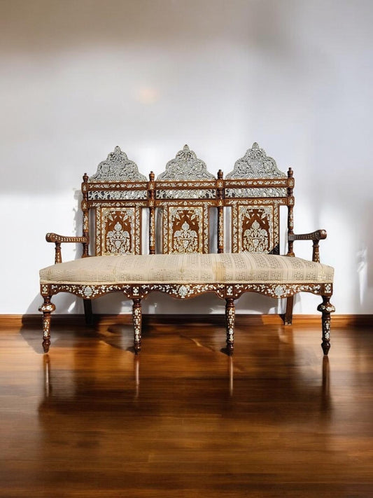 An ornate, antique wooden bench with white shell (mother of pearl) inlays and carved lattice work, positioned in a room with a subtly lit background.