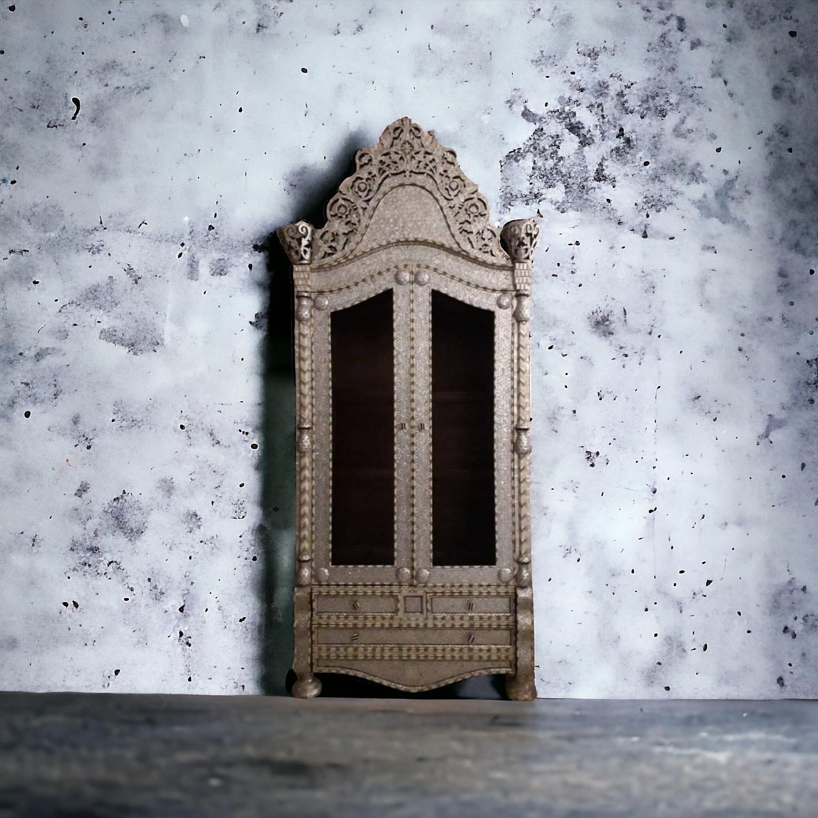 Antique wooden armoire with mother-of-pearl and bone inlay, with a marble top, against a textured wall.