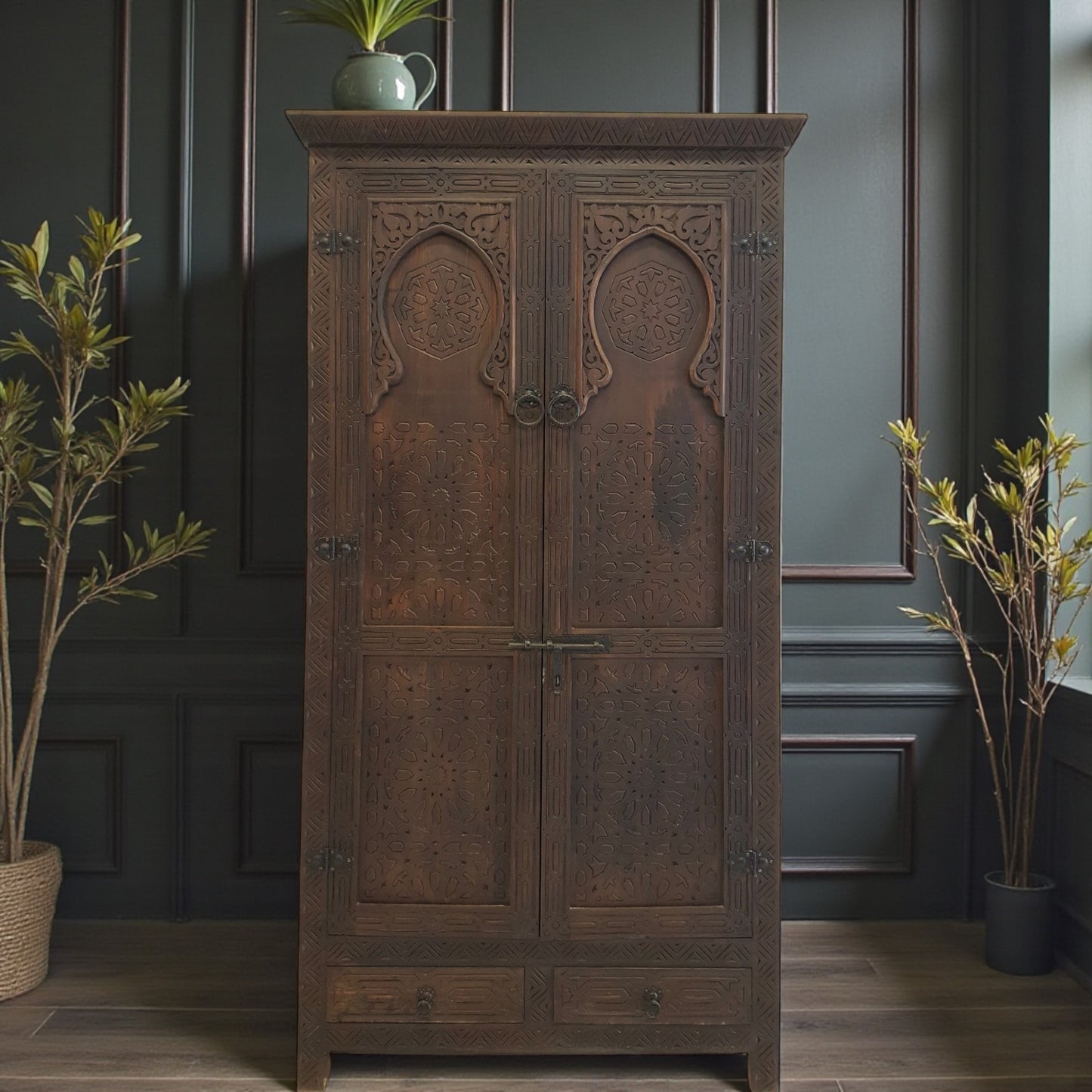 Wooden cabinet with decorative carvings in a room with plants and dark walls.