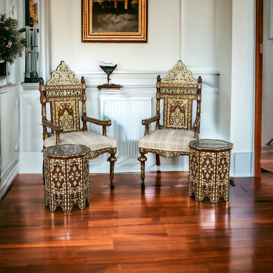 Two antique Syrian mother of pearl inlay chairs with white cushions on a wooden floor, with intricate carvings and a decorative ottoman behind them.