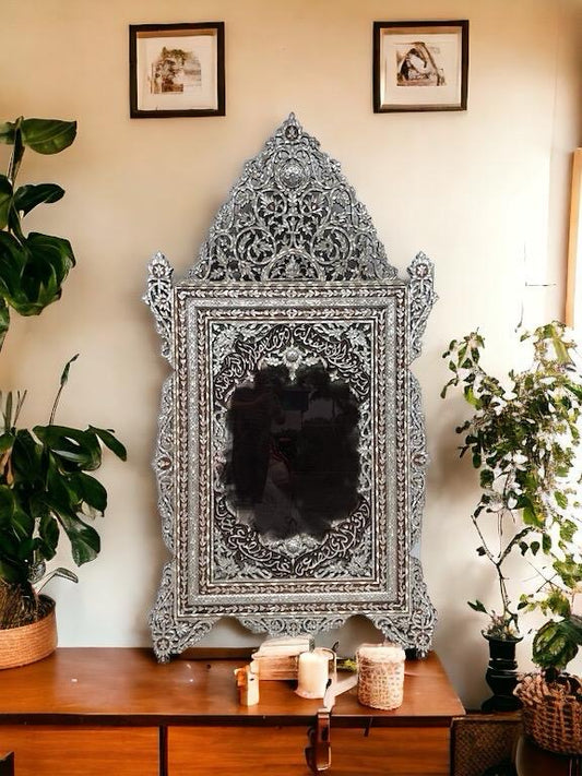 A large vintage Syrian mother of pearl inlay mirror with intricate designs, placed on a wooden surface in a home setting, decorated with candles and plants around it.