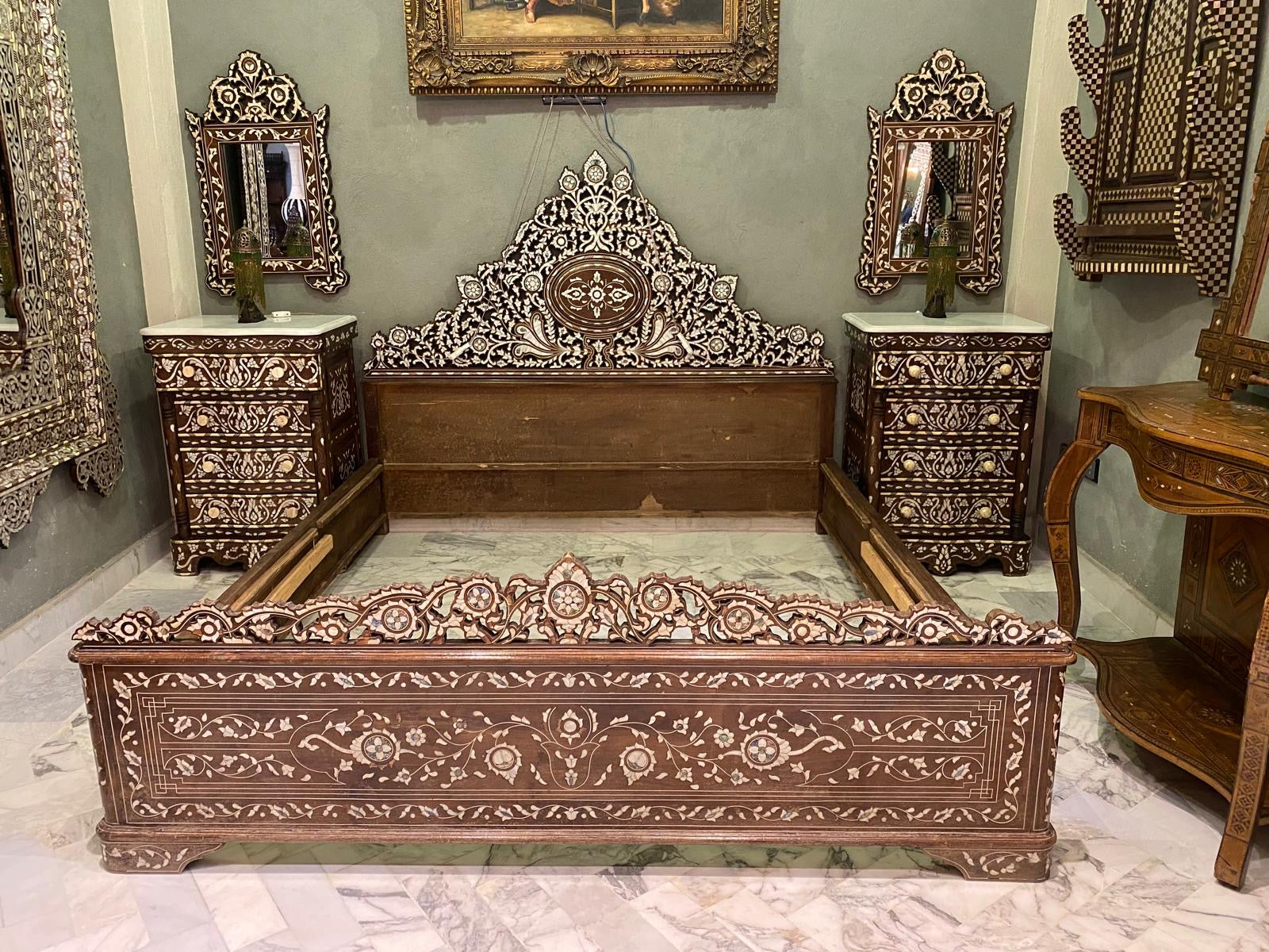 A vintage Levantine royal bed with mother of pearl inlays, accompanied by two matching nightstands, all displayed against a green background with decorative mirrors and furniture.
