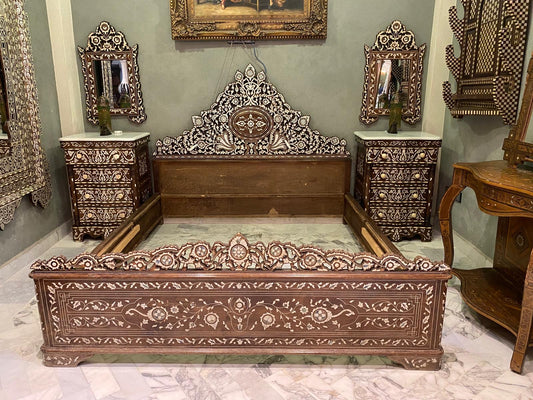 A vintage Levantine royal bed with mother of pearl inlays, accompanied by two matching nightstands, all displayed against a green background with decorative mirrors and furniture.