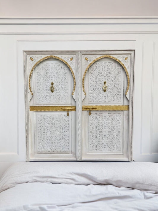 A white, vintage carved cedar double door headboard bed with gold brass knockers and intricate geometric carvings, positioned in a bedroom setting.